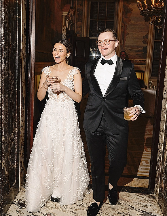 Reception entrance as newlyweds walk through an indoor doorway, bride in lace dress and groom in tuxedo holding cocktail glasses under a chandelier