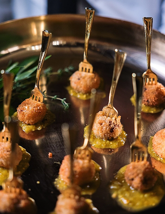 Wedding appetizers and passed hors d'oeuvres on a metal tray with gold cocktail forks, bite-sized croquettes, green sauce, and herb garnish in a blurred reception setting