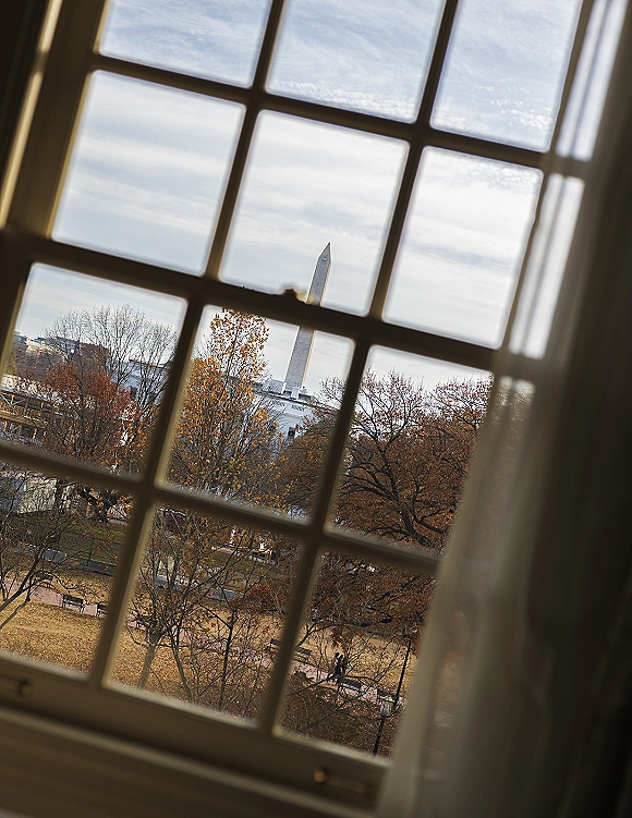 Window view from a bridal suite, framed by window panes, showing park lawns, paths, benches, autumn trees, and a distant obelisk monument.