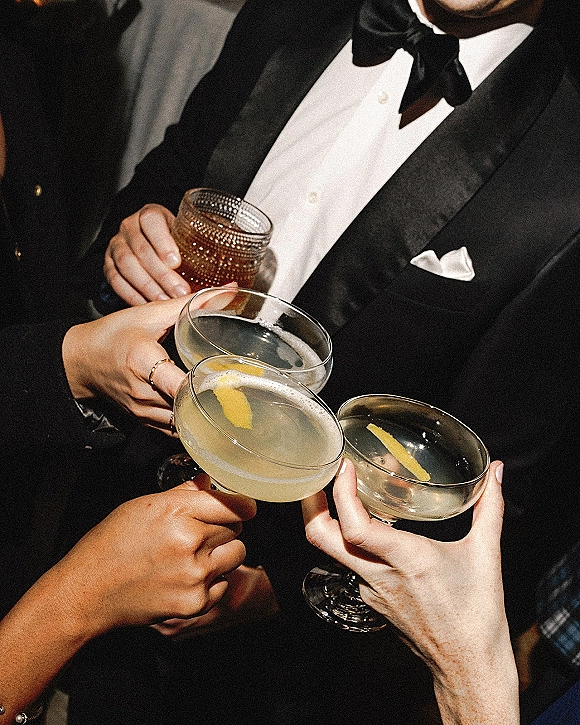 Wedding toast with champagne coupe glasses clinking, lemon twist garnish and tuxedo bow tie visible in dim reception light with blurred guests