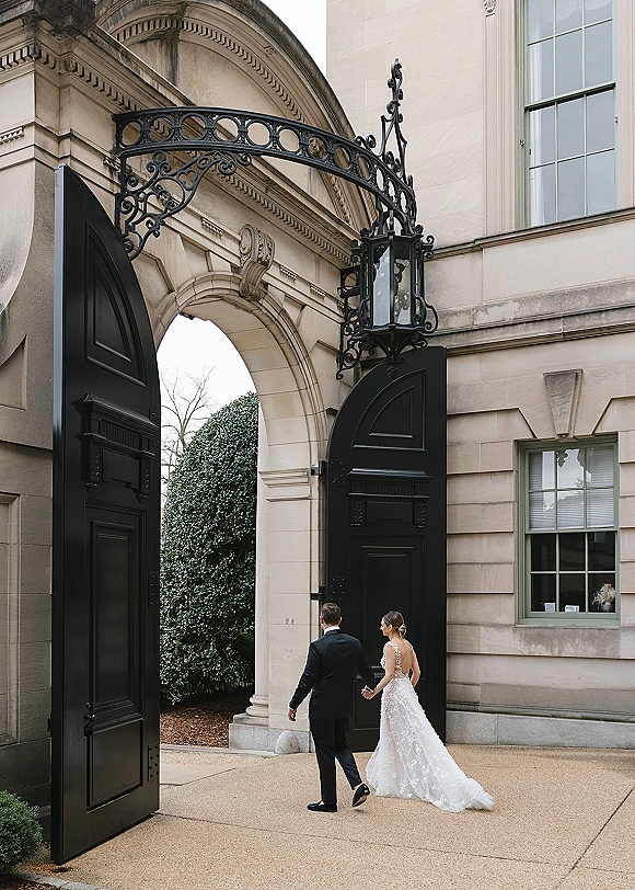 Couple portrait of bride and groom holding hands, walking away toward a stone archway and black doors, her lace train flowing