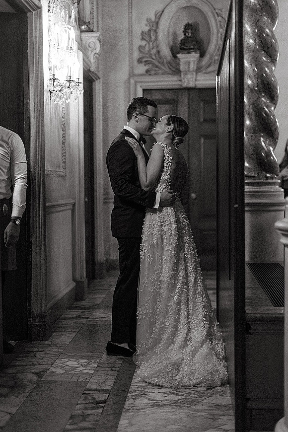 Wedding kiss portrait of bride in a lace applique gown and groom in glasses beneath a chandelier in an ornate marble hallway