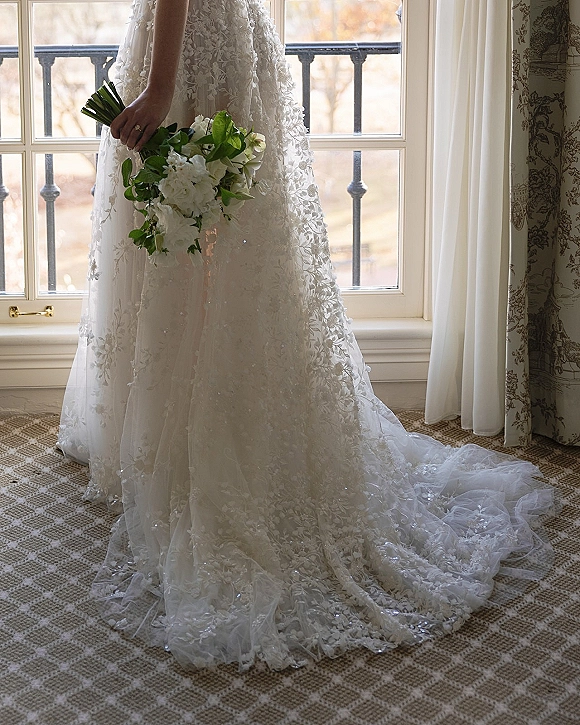 Wedding dress with lace wedding dress appliqué and long train, bride holding white and green bouquet by a large window and balcony railing