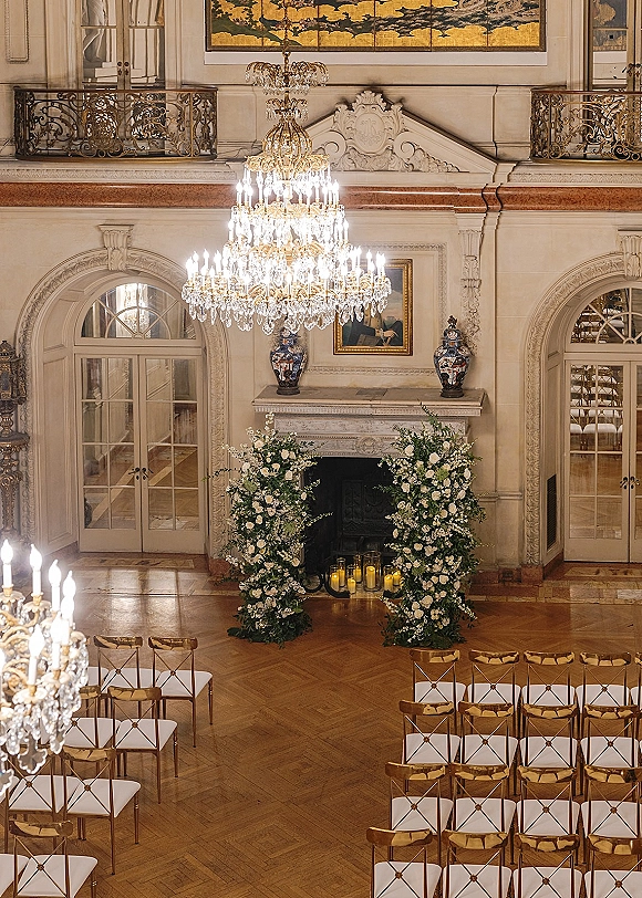 Indoor ceremony setup with a floral ceremony arch framing the fireplace, candlelit aisle, and gold chairs beneath a crystal chandelier