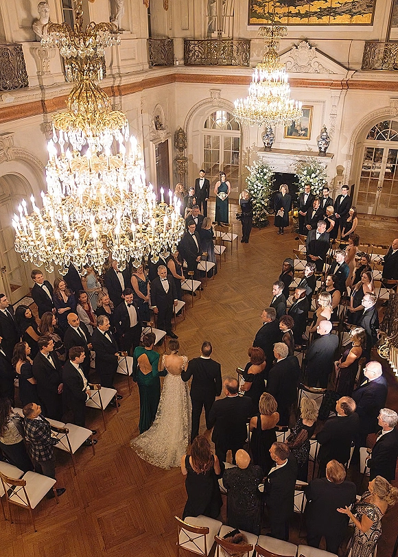 Wedding ceremony in a grand ballroom with crystal chandeliers as the bride walks down the aisle toward a floral arch and candles