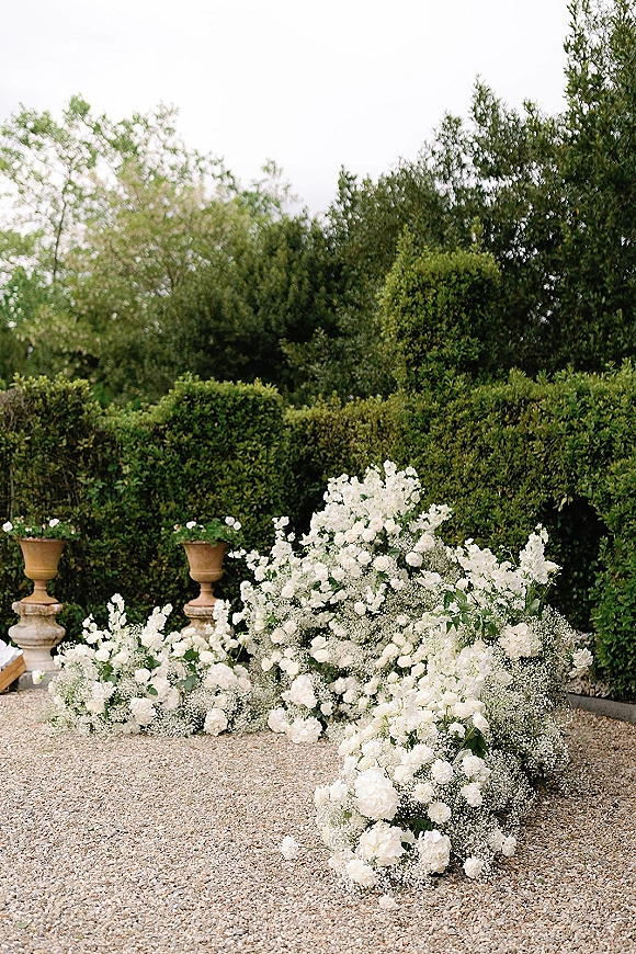 Ceremony floral arrangements of white wedding aisle flowers with baby’s breath and greenery in stone urns on a gravel garden path