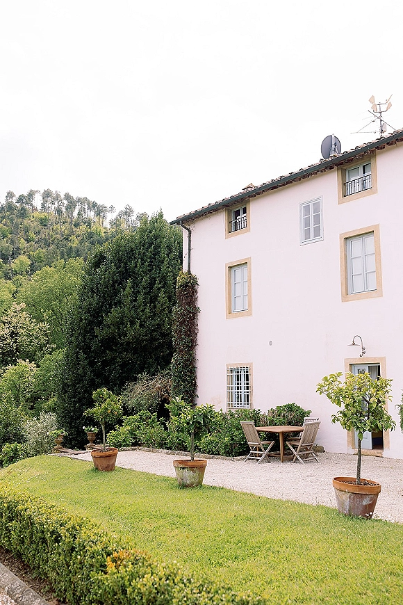Garden patio setting with an outdoor patio table set for two, surrounded by terracotta planters, potted trees, and a white stucco house backdrop
