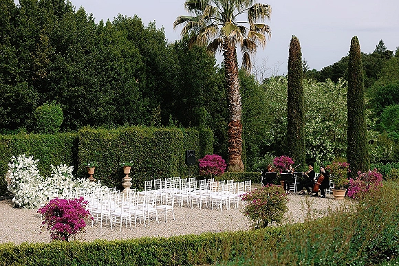 Ceremony setup with outdoor garden ceremony seating, white chairs lining a gravel aisle, string quartet and urn planters in a formal garden
