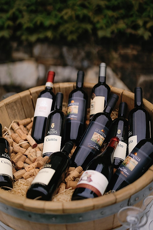 Wine display at a wedding wine station with bottles and corks in a wooden barrel tub filled with straw, set outdoors by greenery and a stone wall