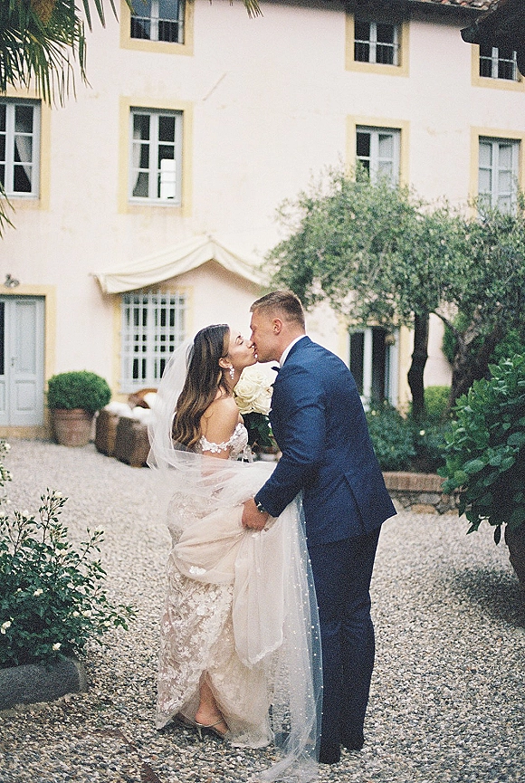 Wedding kiss portrait of bride and groom kissing, bride holding white roses bouquet with veil in a villa courtyard with greenery