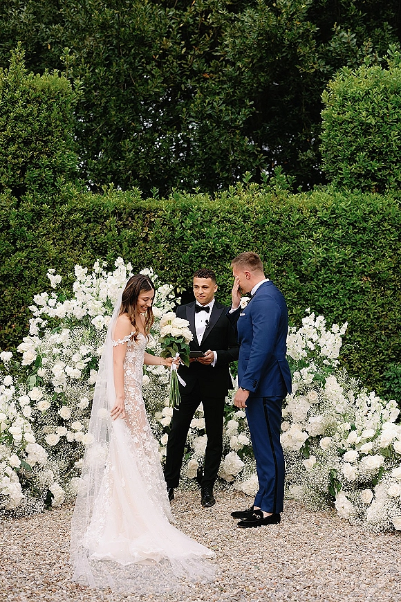 Wedding ceremony moment as bride and groom at altar exchange vows, her white bouquet and veil framed by garden hedges and floral backdrop