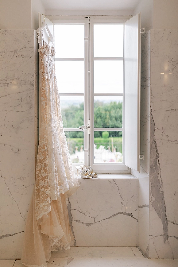 Wedding dress, lace wedding dress on a hanger by a window with marble wall, ivory lace bodice and bridal heels on the sill
