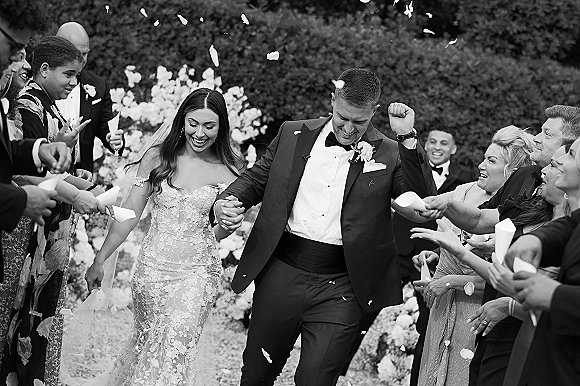Wedding recessional as bride and groom walk the aisle holding hands while guests toss confetti petals along a garden walkway with hedges
