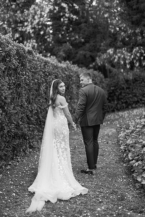 Couple portrait of bride and groom holding hands, walking away on a garden path, her long veil trailing over a lace dress train