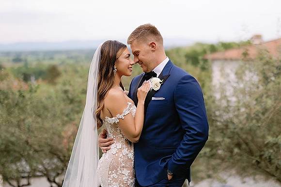 Couple portrait of bride and groom embrace with forehead touch, her long veil and lace off-shoulder gown against greenery and stone building
