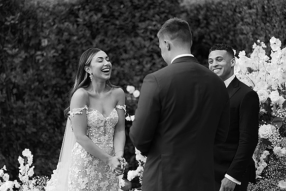 Wedding vows as bride laughs at altar beside groom in black suit, wearing a strapless lace dress and veil, framed by garden hedge and roses