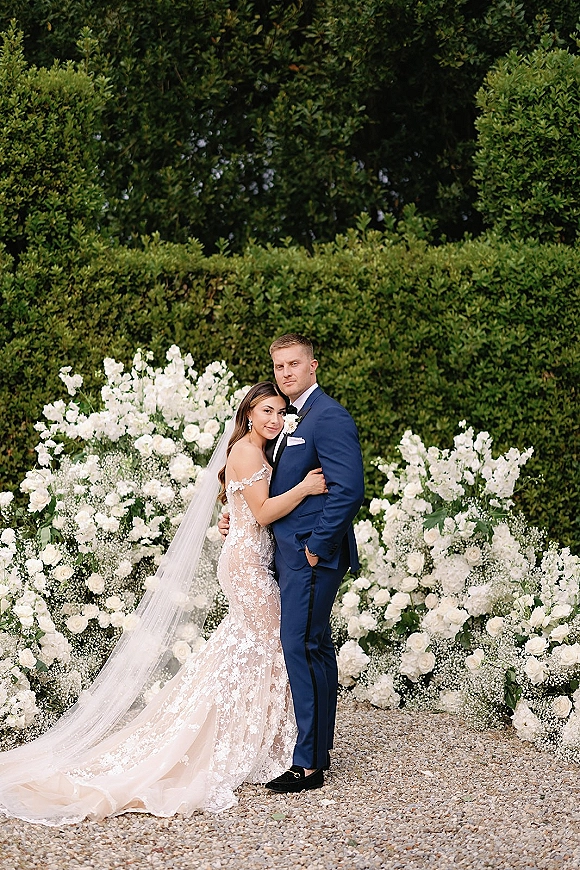 Couple portrait of bride and groom embrace, her long veil trailing over gravel path beside white rose florals and green hedge backdrop