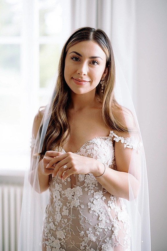 Bridal portrait of a bride in an off the shoulder lace wedding dress, veil over shoulders, lit by window light through white curtains