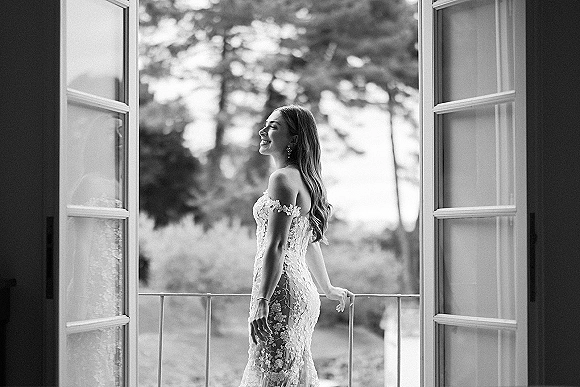 Bridal portrait of a smiling bride in an off-the-shoulder lace wedding dress, side profile by open French doors to a balcony view