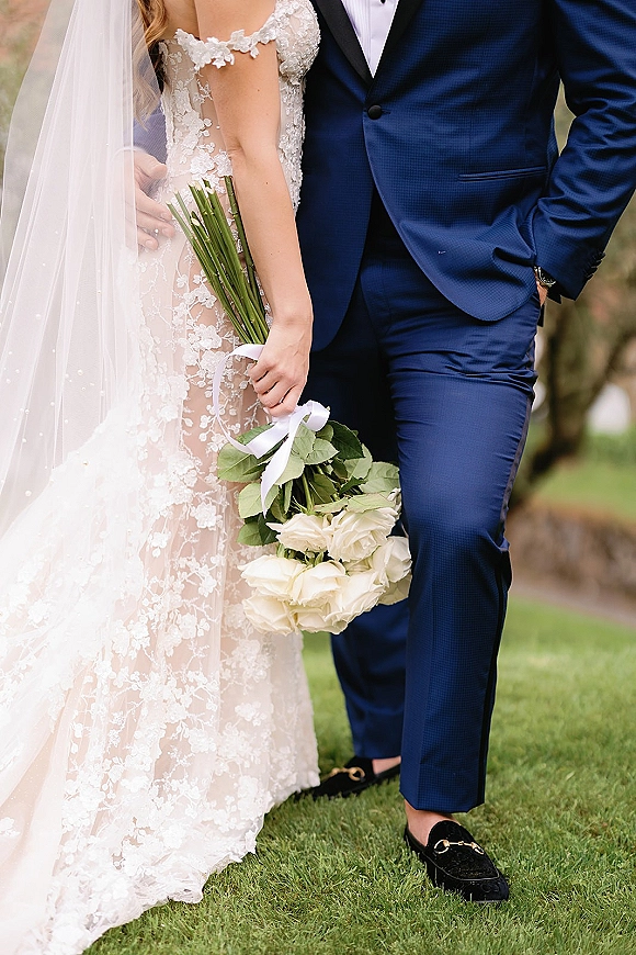 Couple portrait of bride and groom torso portrait, bride in lace gown and veil holding a white rose bouquet with ribbon on a lawn with trees