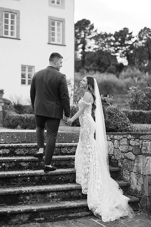 Couple portrait of bride and groom holding hands, walking up stone steps beside a stone wall, her long veil trailing behind