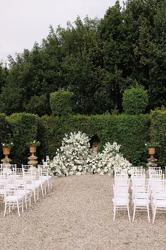 Ceremony setup for an outdoor wedding ceremony with white chiavari chairs lining a gravel aisle, white florals and stone urns by a hedge wall