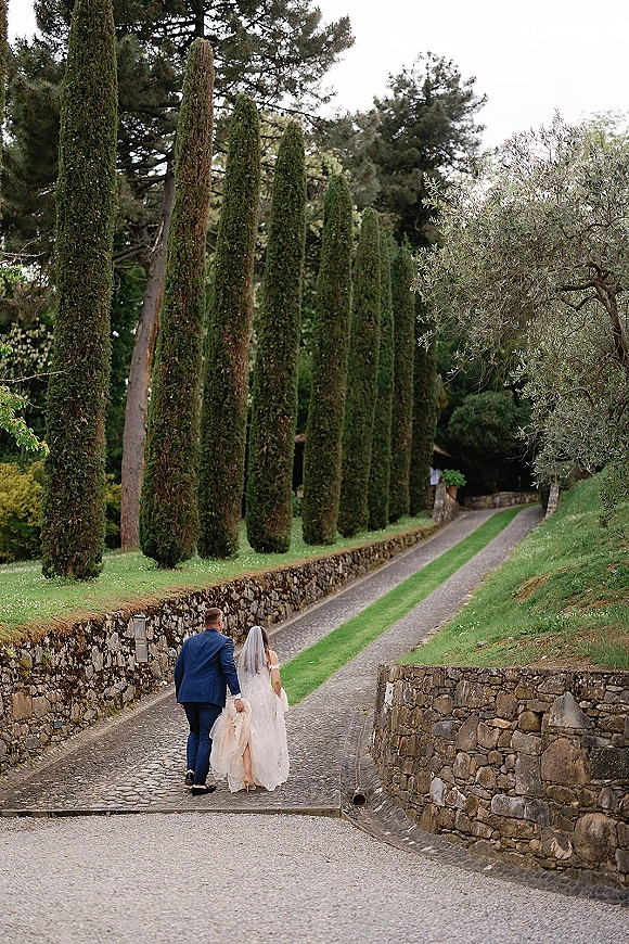 Couple walking portrait of bride and groom walking away, veil flowing as she lifts her train along a tree-lined cobblestone drive and stone walls