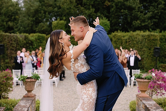 Wedding kiss portrait of bride and groom kissing in a garden aisle, her lace veil flowing as guests watch near white chairs and potted flowers