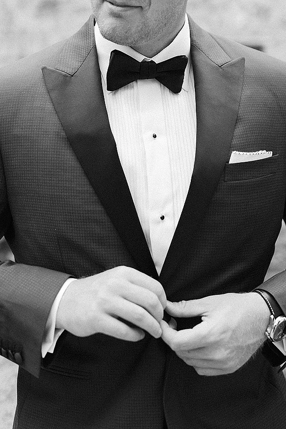 Groom portrait in a black tie groom tuxedo, showing bow tie, satin lapel, pocket square, and wristwatch against blurred outdoors