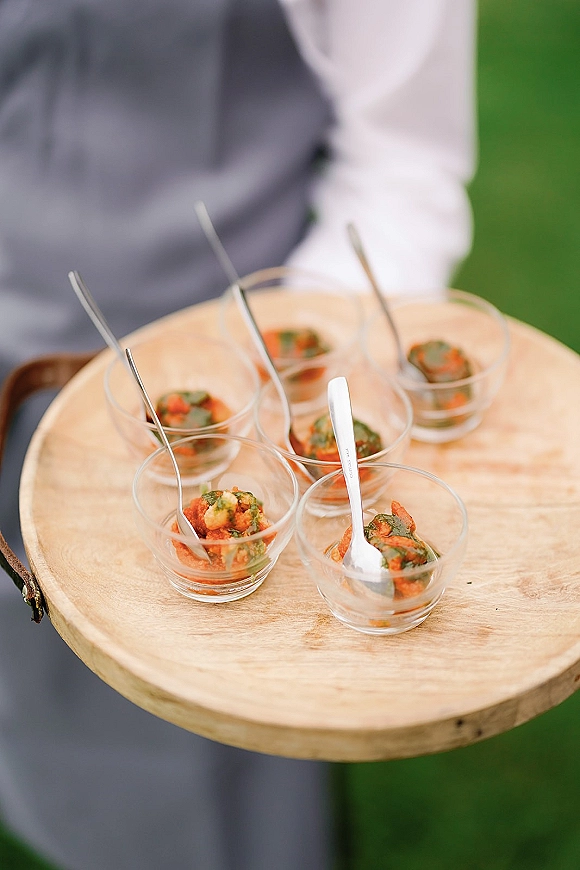 Wedding passed appetizers served in glass cups with tasting spoons on a wood tray, held by a vested server over a green lawn