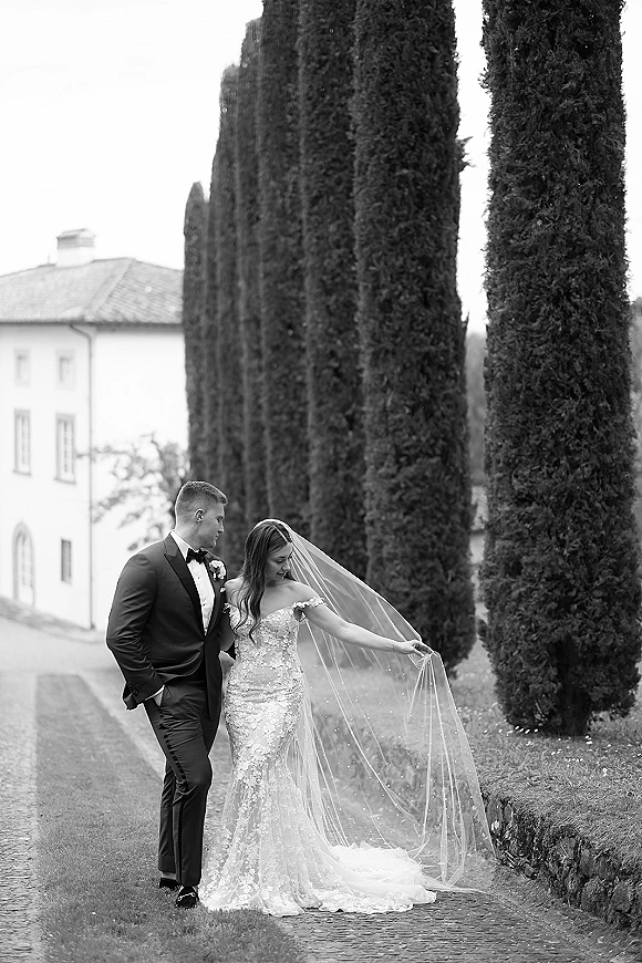 Couple portrait, black and white wedding portrait of bride holding her veil beside groom in tuxedo on a cobblestone path by cypress trees and villa