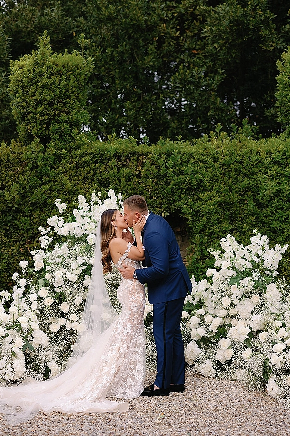Wedding kiss as bride in lace dress and long veil cups groom’s face, his blue suit framed by garden hedges and gravel path
