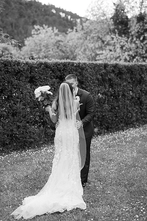 Wedding kiss portrait of bride and groom kissing, her long veil and white bouquet against a hedge garden with distant hills beyond