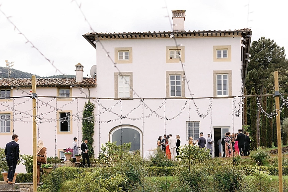 Cocktail hour with wedding guests mingling under string lights, holding drinks on a villa lawn by an arched doorway and hedges