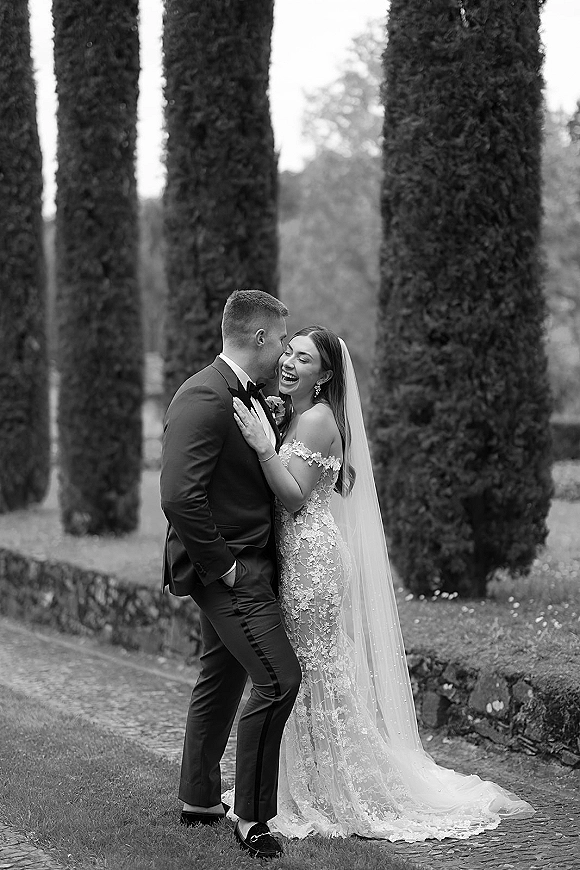 Couple portrait in black and white of bride and groom laughing as he kisses her cheek, her veil draped in a garden by a stone wall