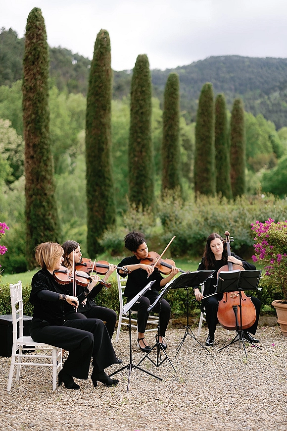 Wedding string quartet ceremony musicians performing with violins and cello beside white chairs on a garden gravel patio with cypress trees