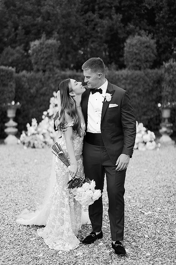 Couple portrait in a black and white wedding portrait style, bride and groom embrace on a gravel path by garden hedges with veil and bouquet
