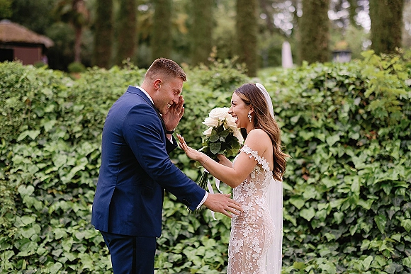 Wedding first look as the bride taps the groom’s shoulder, holding a white rose bouquet by an ivy wall in a garden setting