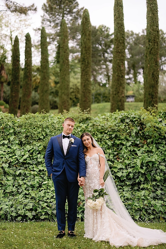 Couple portrait of bride and groom holding hands, her veil and bouquet beside his navy tuxedo, posed before an ivy wall and cypress trees