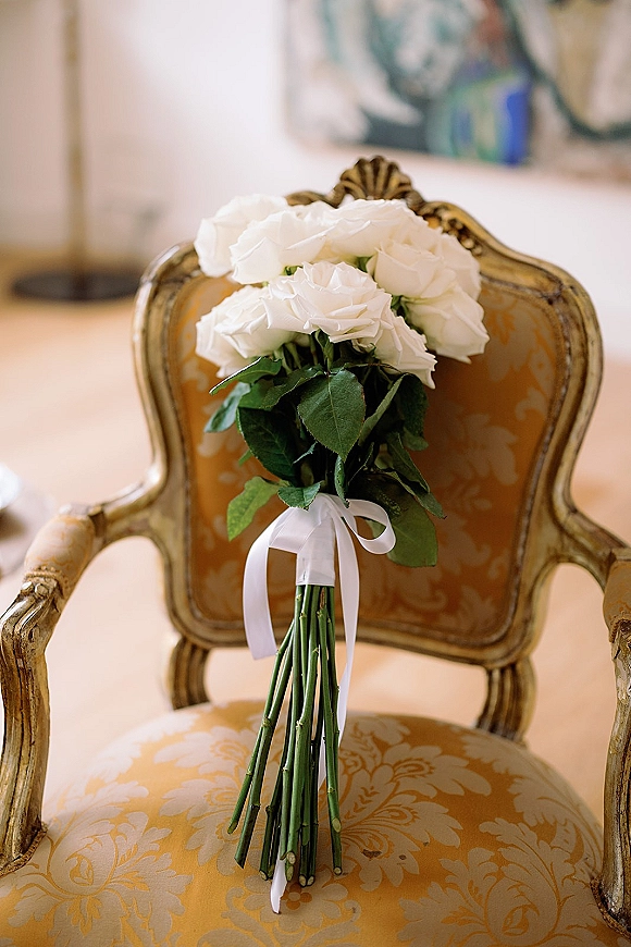 Bridal bouquet of white roses with greenery and a white ribbon, resting on a vintage upholstered chair in an indoor room with framed art