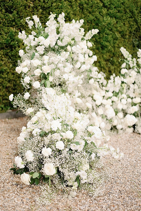 Ceremony aisle flowers in white roses and baby's breath with greenery, arranged low along a gravel path against a hedge wall backdrop