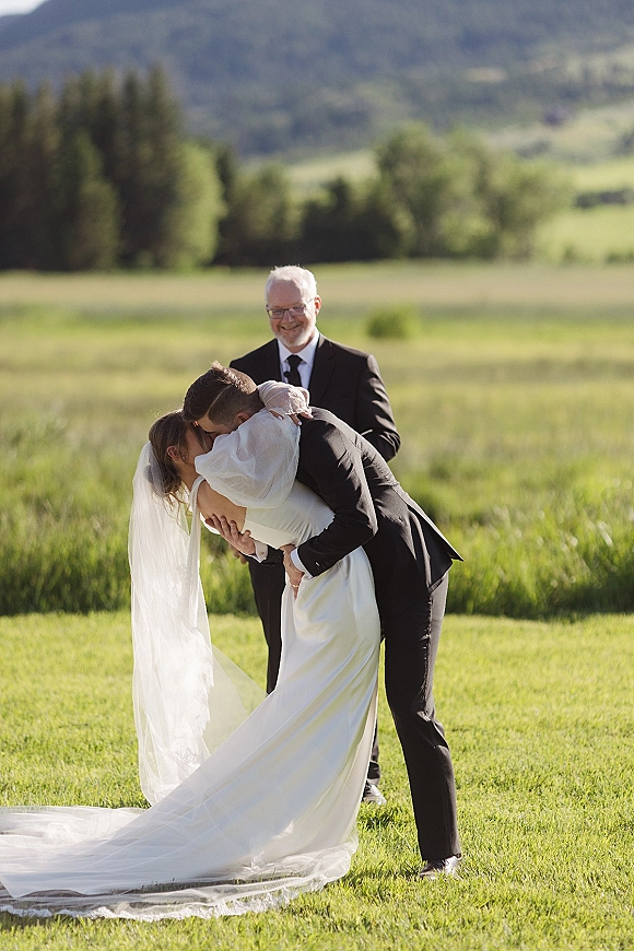 Wedding kiss portrait of groom dipping the bride as they kiss, her long veil flowing, in a grassy field with mountains behind