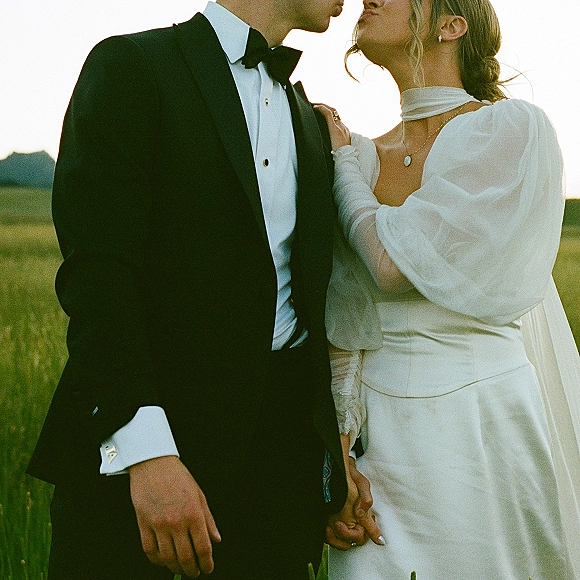 Couple portrait of bride and groom holding hands in a tall grass field at sunset, her veil draped over shoulder and his tuxedo bow tie visible