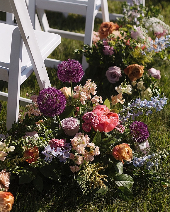 Ceremony aisle flowers with a ground arrangement of roses, peony, allium, snapdragons and delphinium beside white folding chairs on a grass lawn