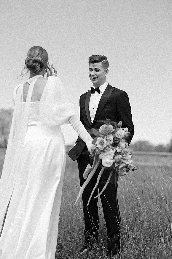 First look moment as bride walks up from behind in long sleeve wedding dress, holding bouquet with ribbon in a meadow field