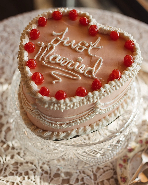 Wedding cake in heart shaped design with pink icing and piped frosting, topped with maraschino cherries on a glass stand over lace tablecloth