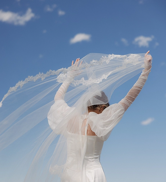 Bridal portrait of a bride with veil, lace edge veil flowing in the wind as she holds it up against a blue sky with clouds