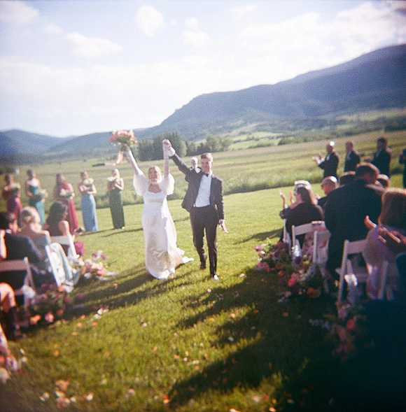 Wedding recessional as bride and groom walk the petal-strewn aisle, bride lifting her bouquet, guests cheering with mountains behind