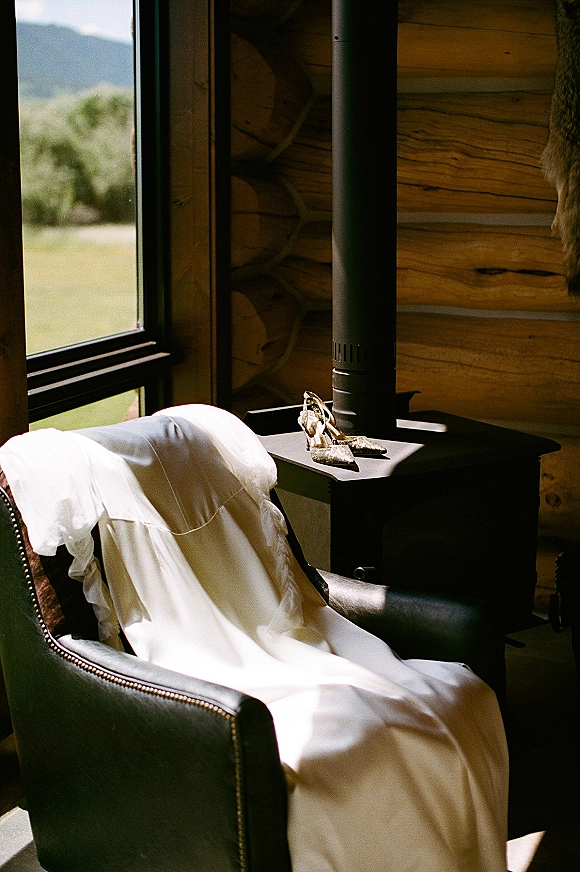 Wedding dress detail with gold bridal heels on an armchair beside a wood stove, lit by window light in a rustic cabin interior