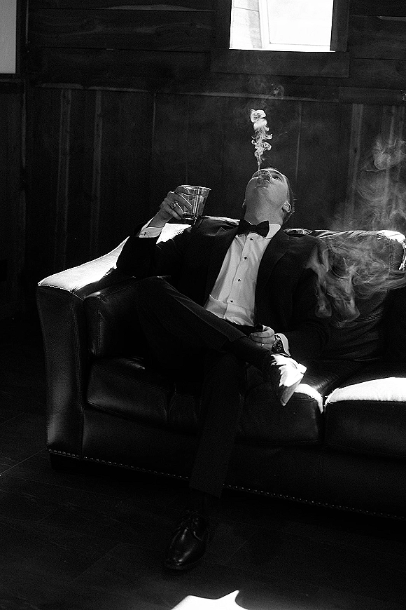 Groom portrait in tuxedo seated on a leather sofa, holding a cigar and whiskey glass, lit by window light against wood paneling